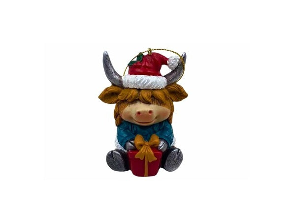 A Highland cow Christmas ornament wearing a red Santa hat, blue outfit, and holding a red gift box with a gold bow, set against a plain white background.